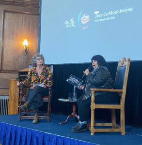 Two women on a stage. On the right, poet Jess Mookherjee, holding microphone and looking surprised. On the left, writer Sarah Jane Butler, listening.