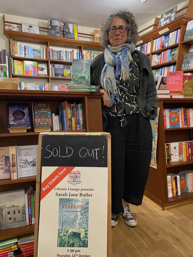 Middle-aged woman in glasses, blue scarf and baggy trousers leaning on a shop counter behind a sign advertising a sold-out literary event for her book, Starling